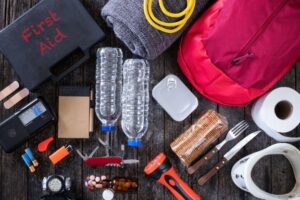Supplies for an emergency preparedness kit are spread across a wooden table. There are water bottles, utility knives, medicine, flashlights, and more.